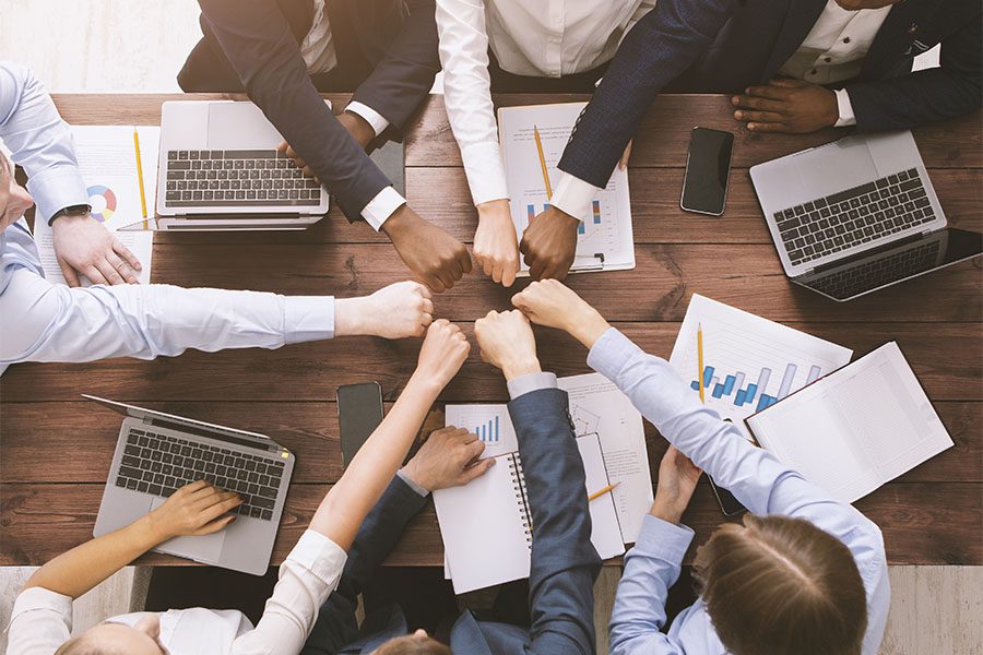 About Our Agency - Group Of Coworkers Sitting At Wooden Table In Office Putting Their Fists Together In Unity